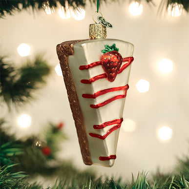 Glass cheesecake slice ornament with red drizzle and a strawberry on top, hanging on a tree.