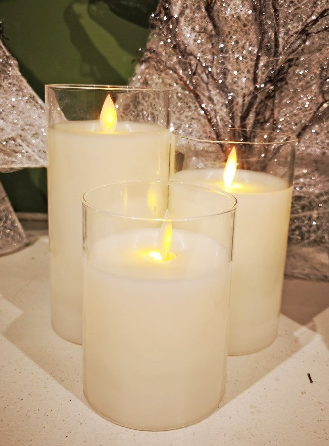 Three cream-colored LED pillar candles of varying heights inside clear glass cylinders.