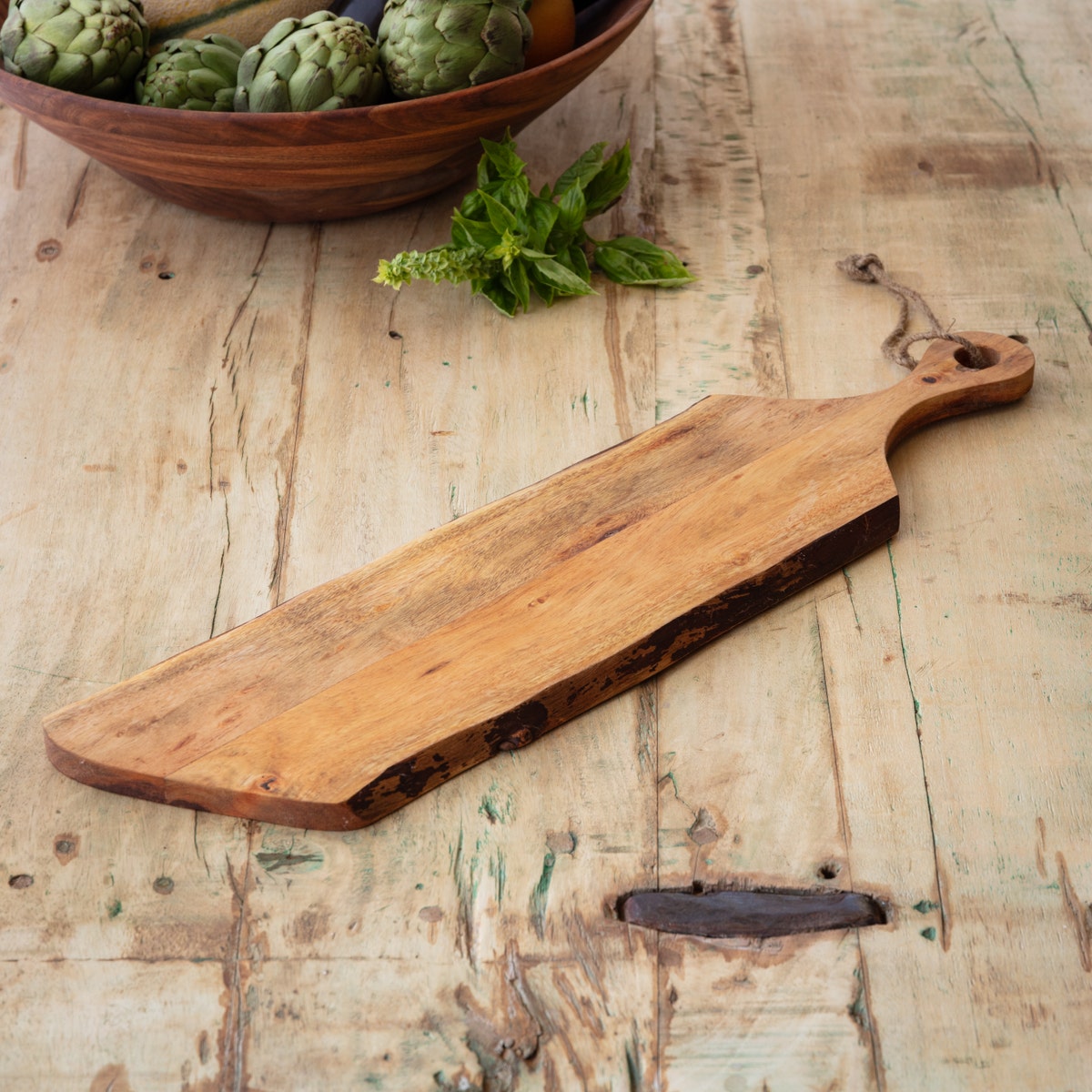 Long wooden live edge baguette board with handle and hanging rope on a rustic table.