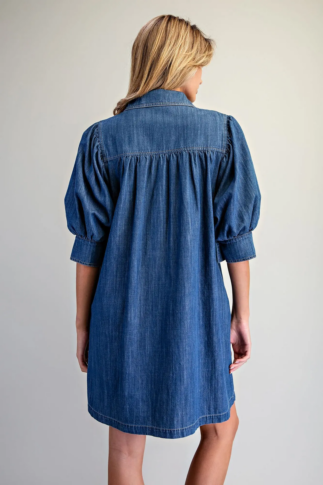 Back view of a blue denim mini dress with puff sleeves and gathered detailing at the yoke.