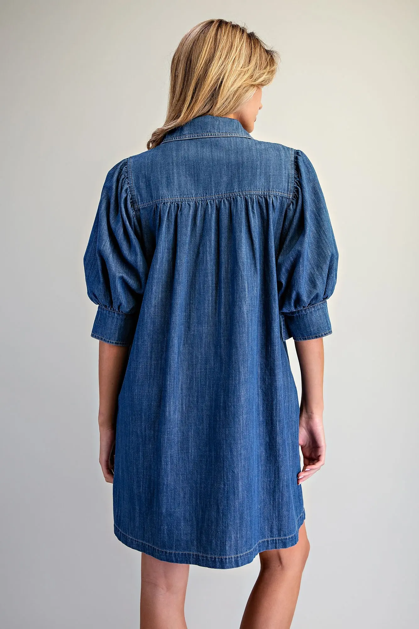 Back view of a blue denim mini dress with puff sleeves and gathered detailing at the yoke.