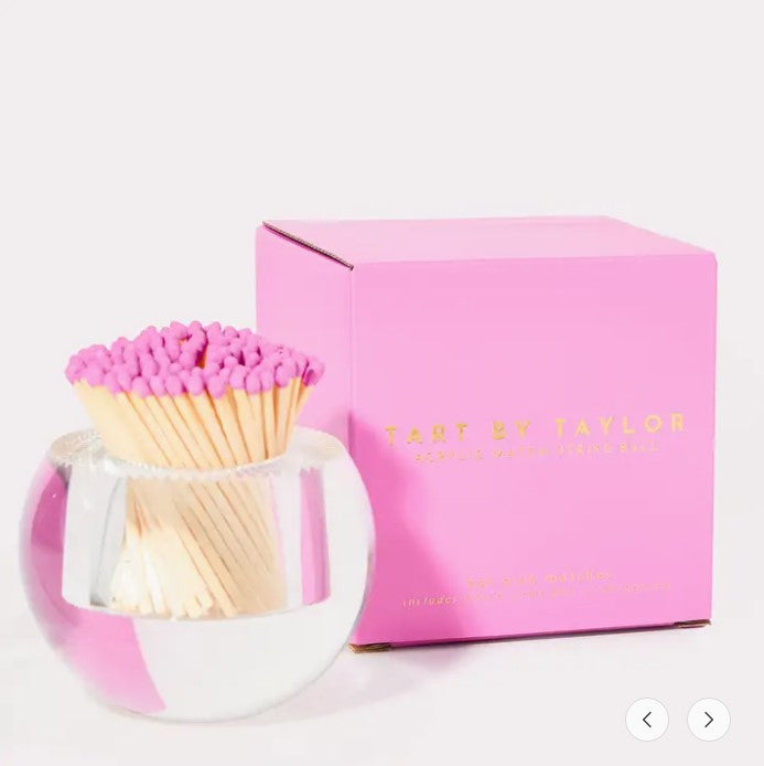 Round clear acrylic match striker with pink-tipped matches next to its pink branded box.