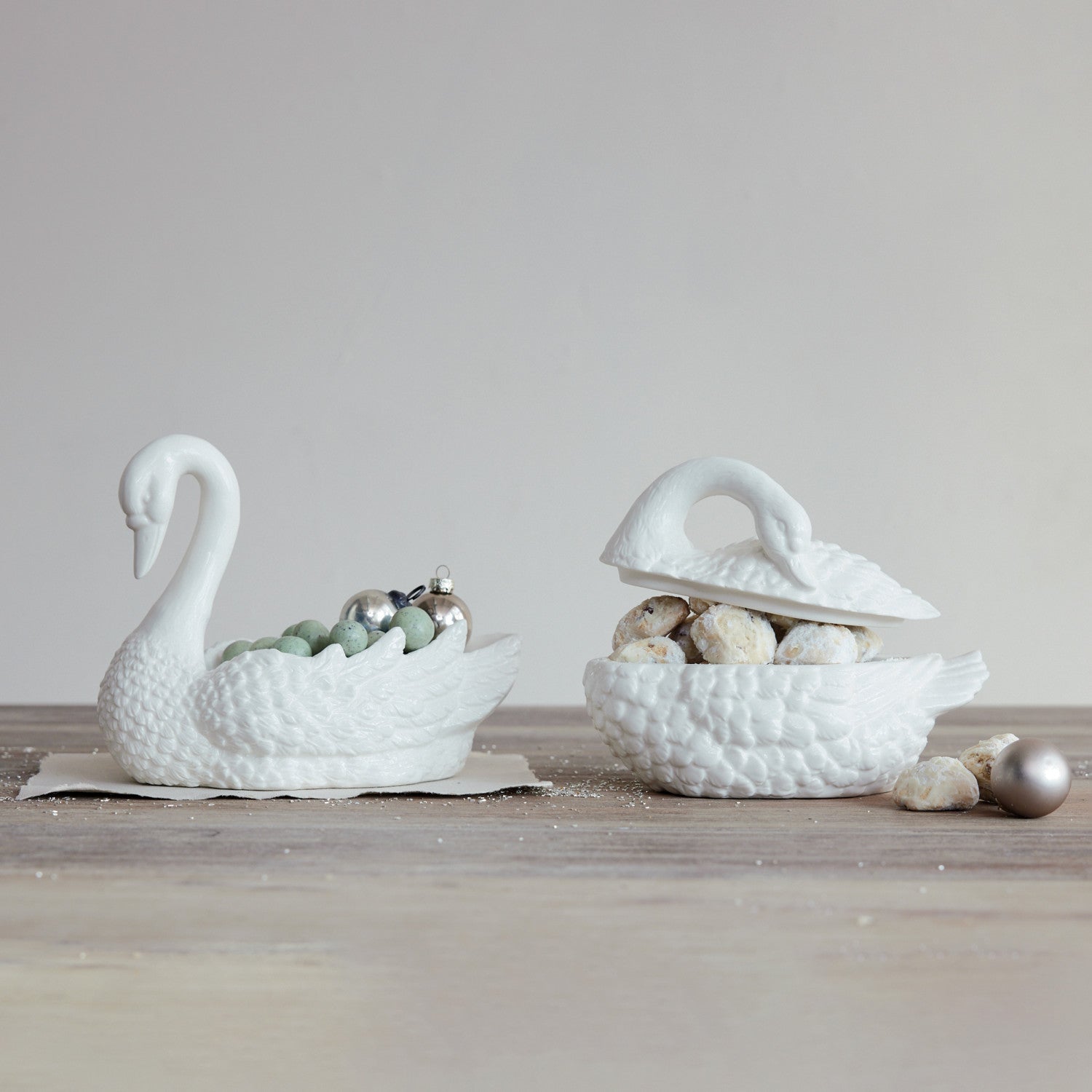 Two white stoneware swan-shaped jars, one open showing cookies and one filled with ornaments.