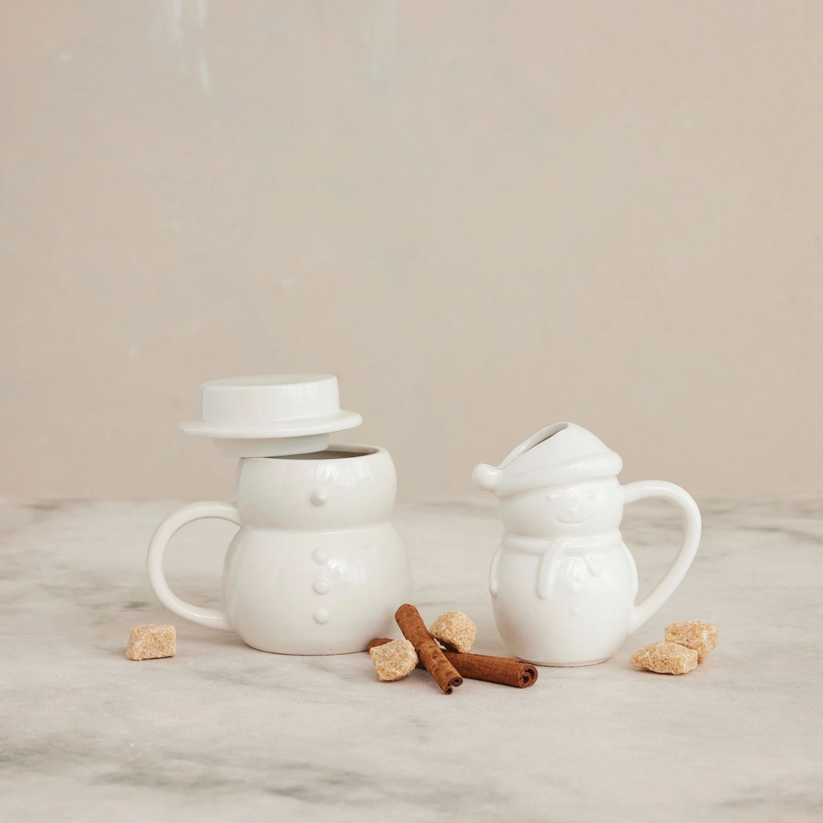 White stoneware snowman mug with a hat-shaped lid and a matching snowman creamer on a marble surface