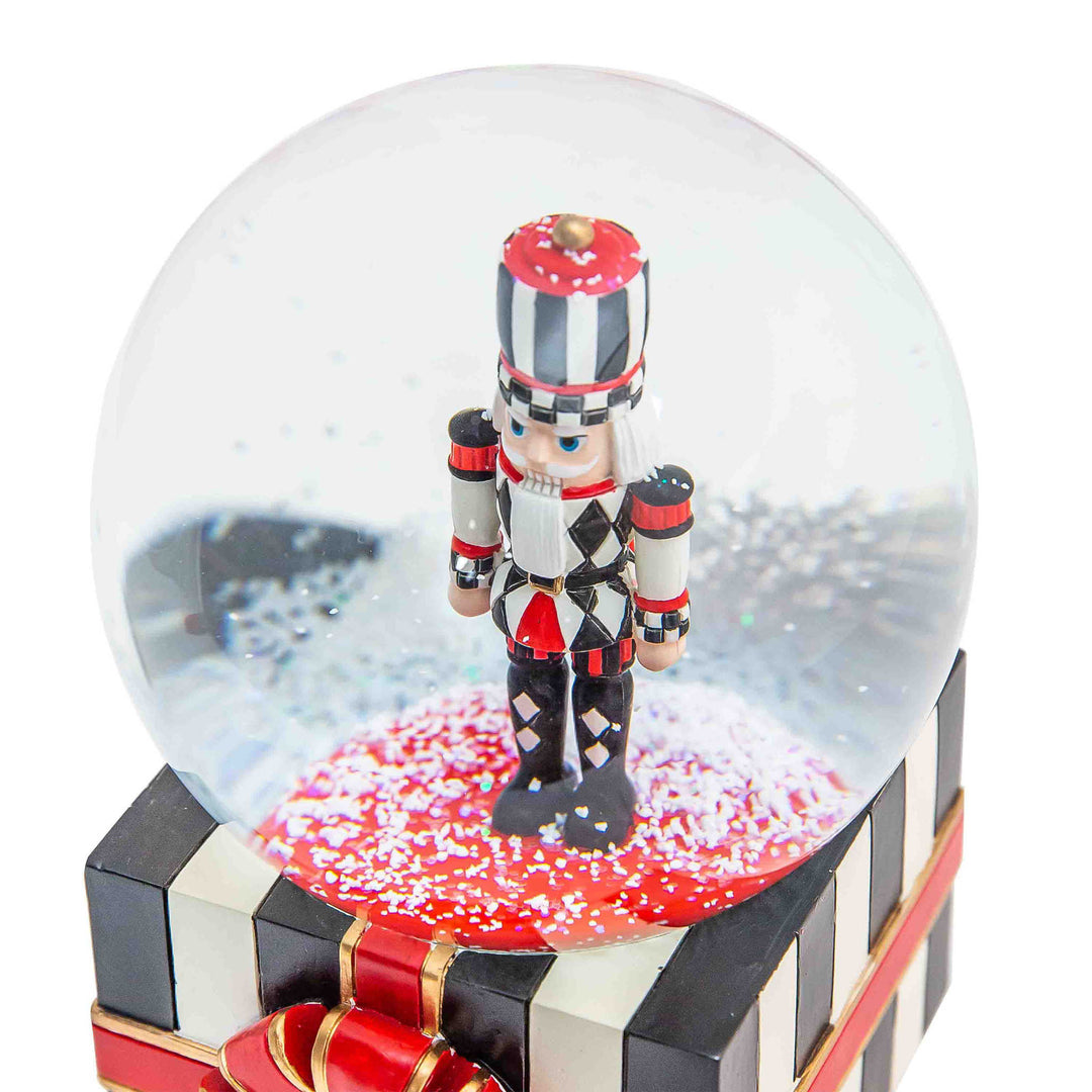 Resin nutcracker snow globe with striped gift box base, red ribbon, and Courtly Check ball pattern.