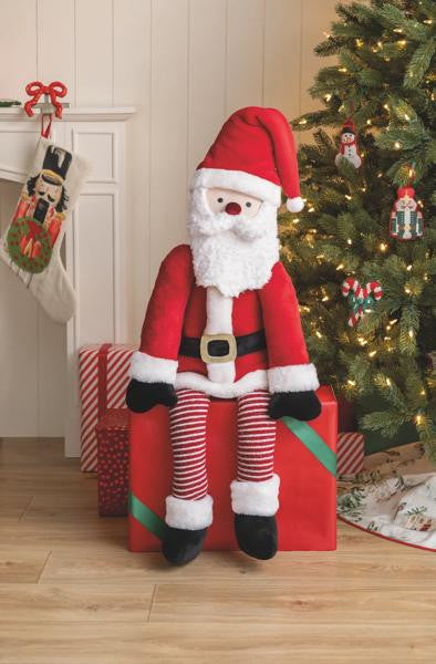 Large plush Santa Claus doll with long striped legs sitting on a red gift box by a Christmas tree.