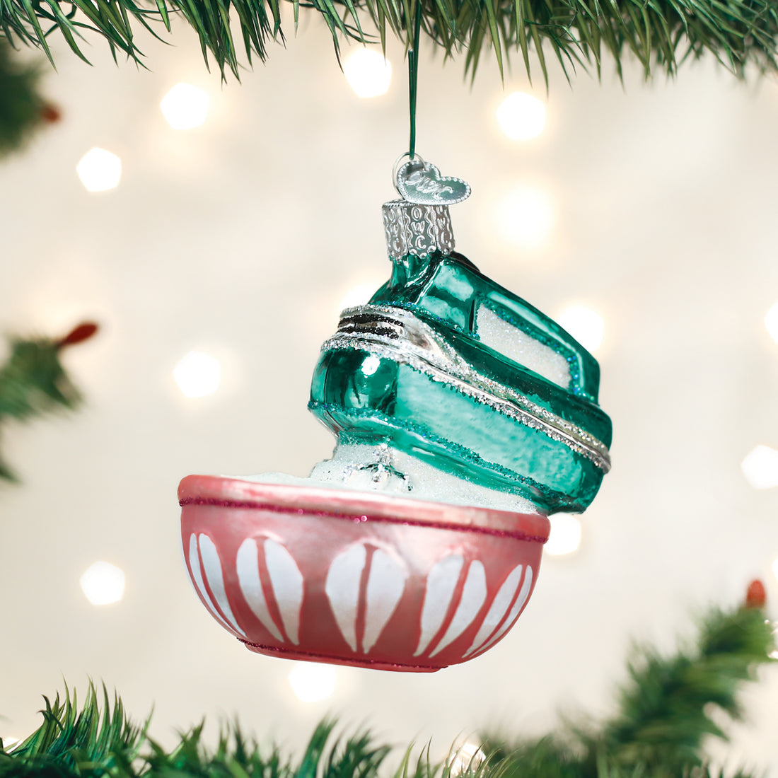 Glass hand mixer ornament with a teal mixer and pink bowl hanging on a Christmas tree branch.
