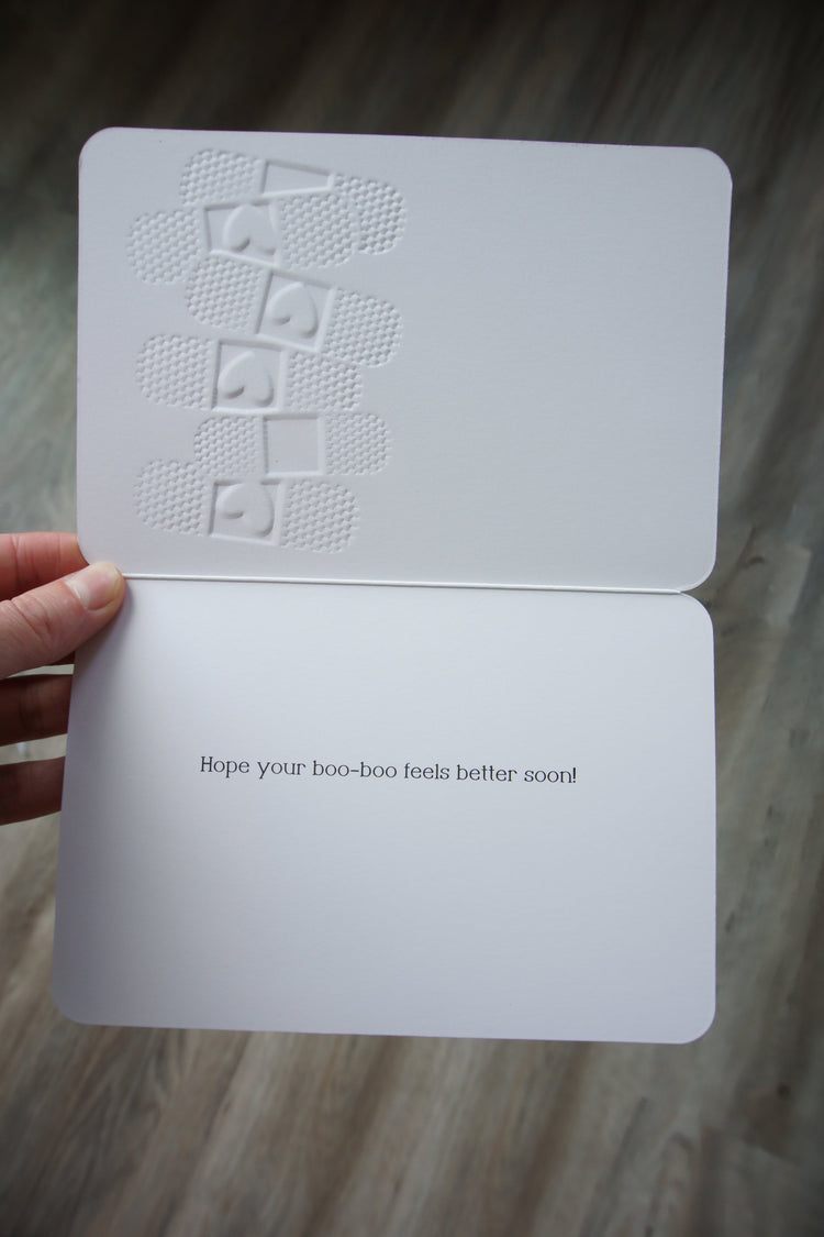 Die-cut get well card shaped like a bandage with embossed texture; outside reads Ouch and inside says Hope your boo-boo feels better soon.