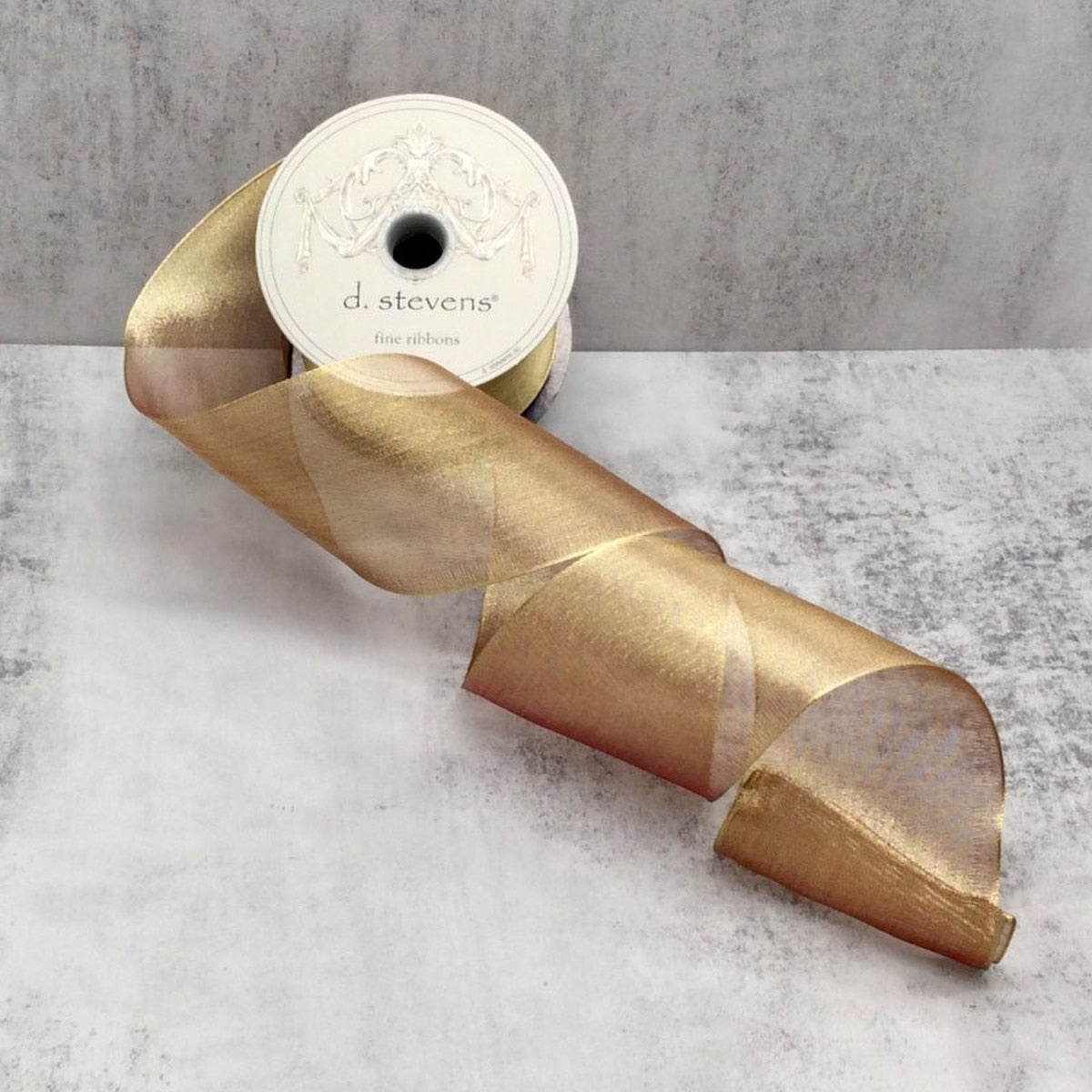 A spool of metallic gold mesh ribbon by d. stevens on a light grey textured surface.