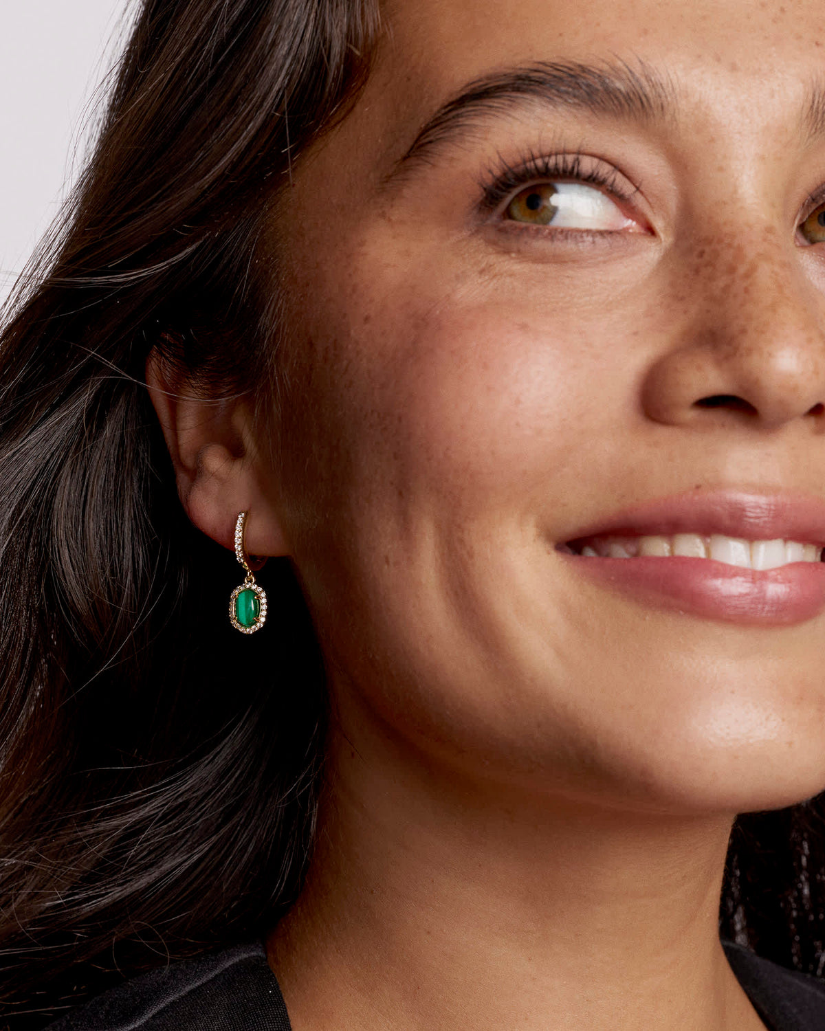 Close-up of a woman wearing a Daphne Crystal Frame Huggie Earring with a green oval charm.