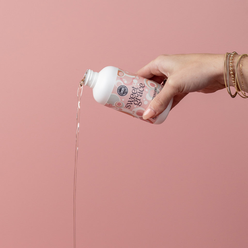 A hand pouring Sweet Grace 6oz Laundry Detergent from a white bottle against a pink background.