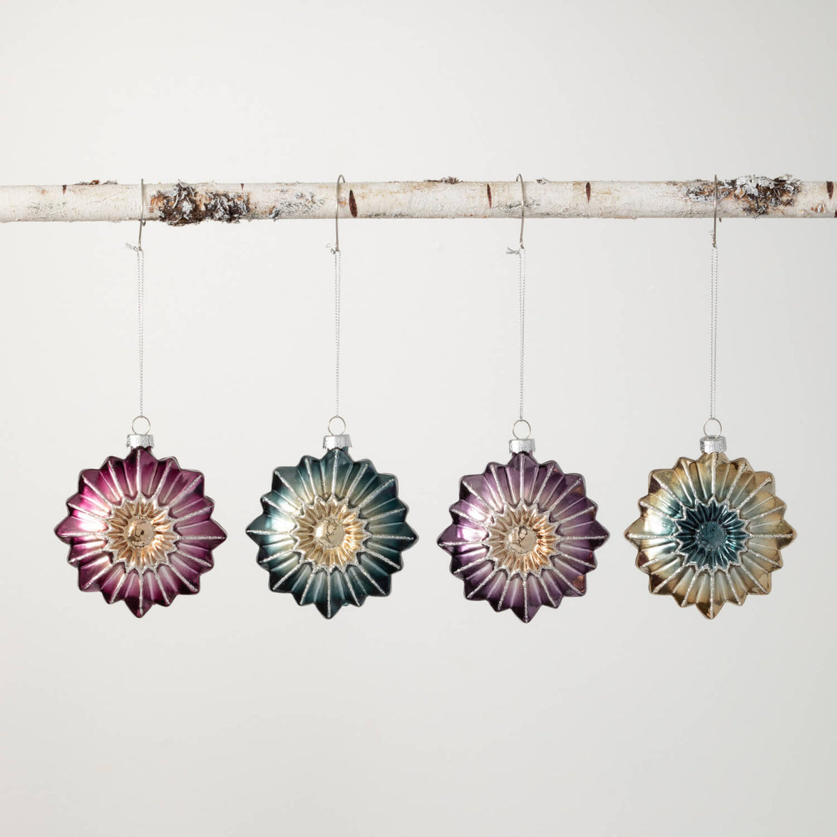 Four star-shaped glass witch eye ornaments in purple, blue, and gold hanging from a birch branch.