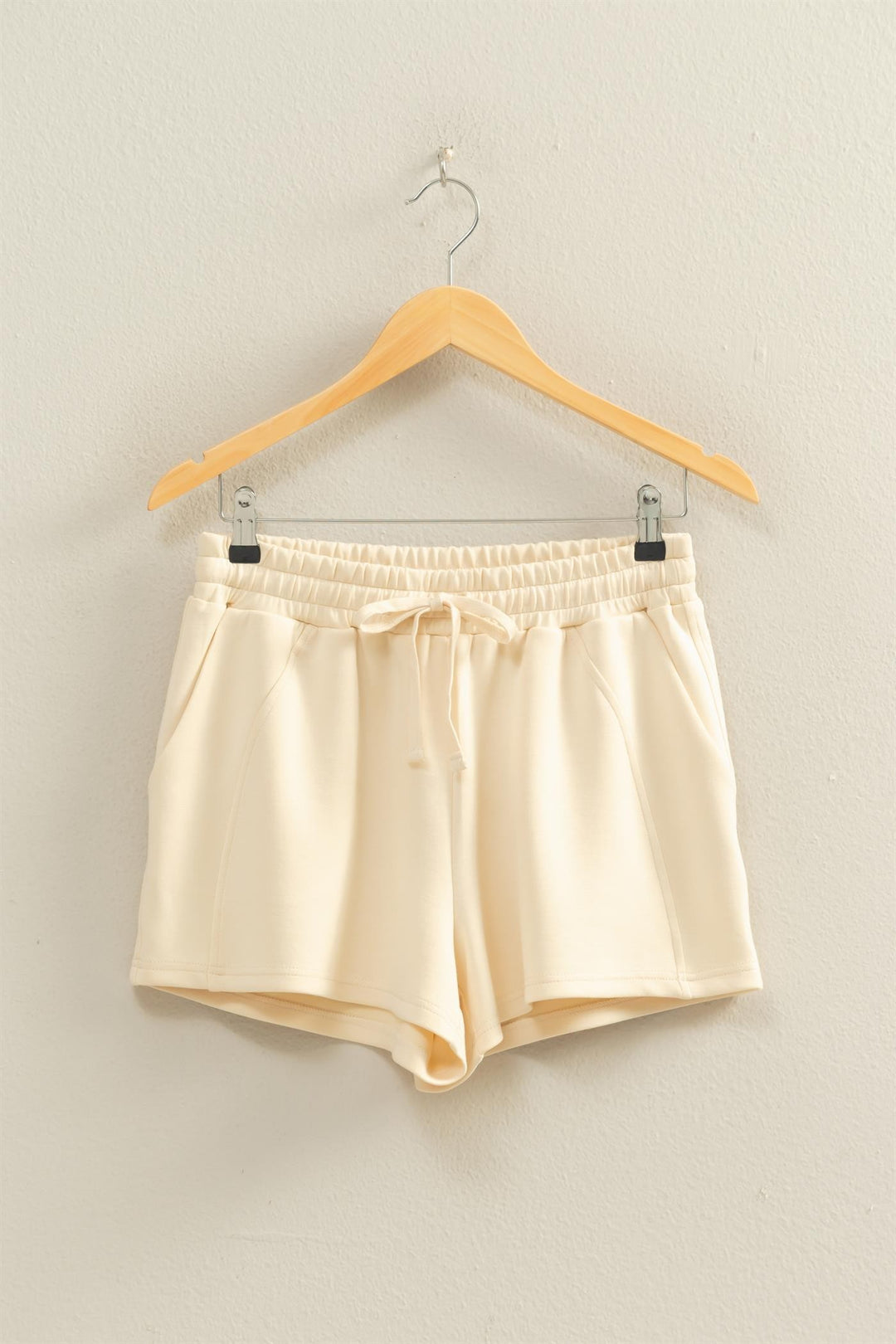 Cream-colored Sylvia drawstring waist shorts hanging on a wooden hanger against a neutral wall.