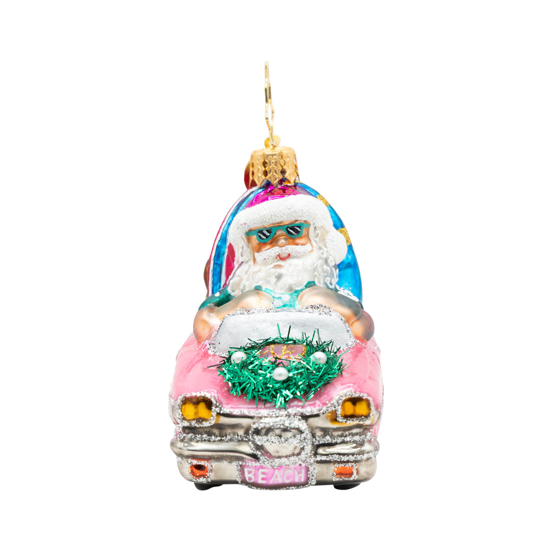 Santa Claus wearing sunglasses driving a pink convertible with a surfboard and a wreath on the hood