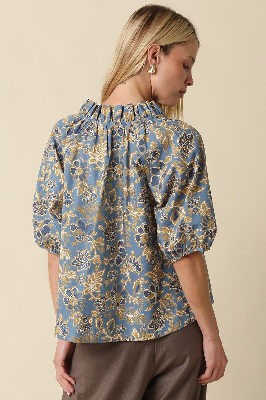 Back view of a woman wearing a blue floral Marina blouse with puff sleeves and a ruffled neckline.