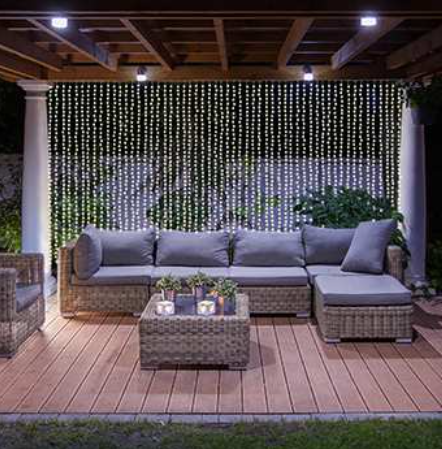 Warm white connectable curtain lights hanging behind a grey wicker patio sofa set at night.
