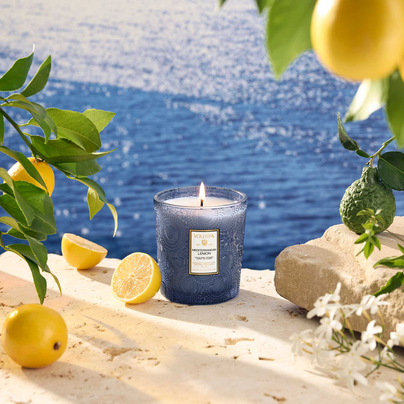 Mediterranean Lemon 9oz. Classic Candle in a blue glass jar on a stone ledge by the sea.