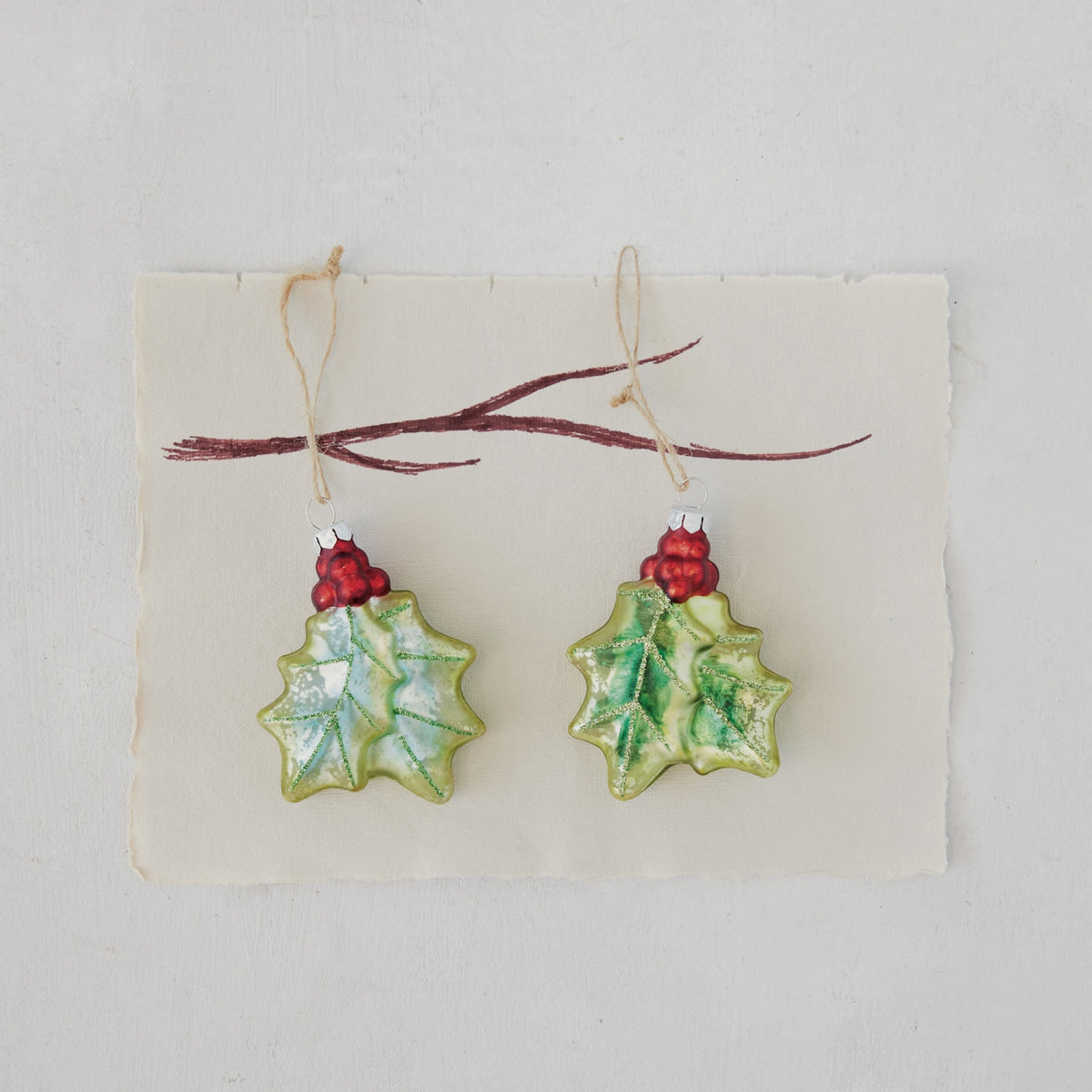 Two hand-painted mercury glass holly ornaments with red berries and glitter on a twine loop.