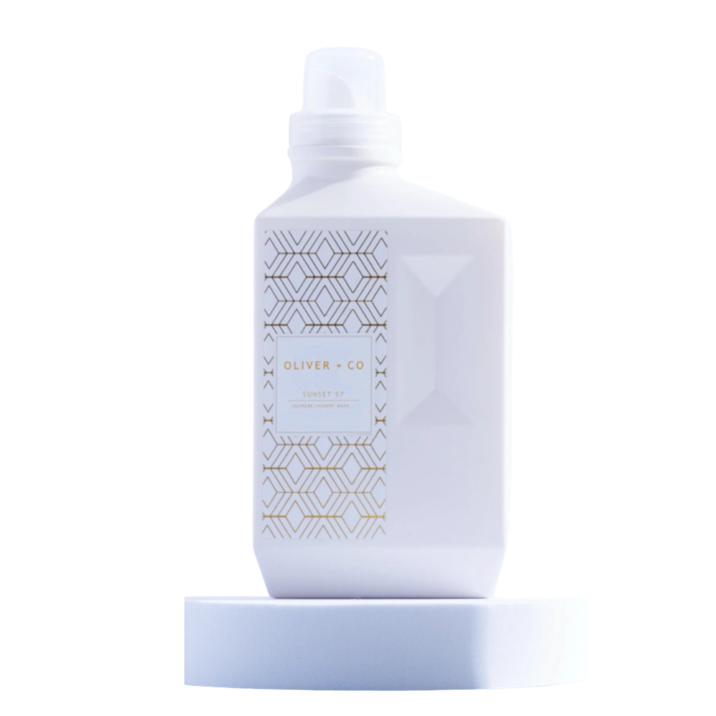 White bottle of Oliver + Co Supreme Laundry Wash with gold geometric patterns on a white pedestal.