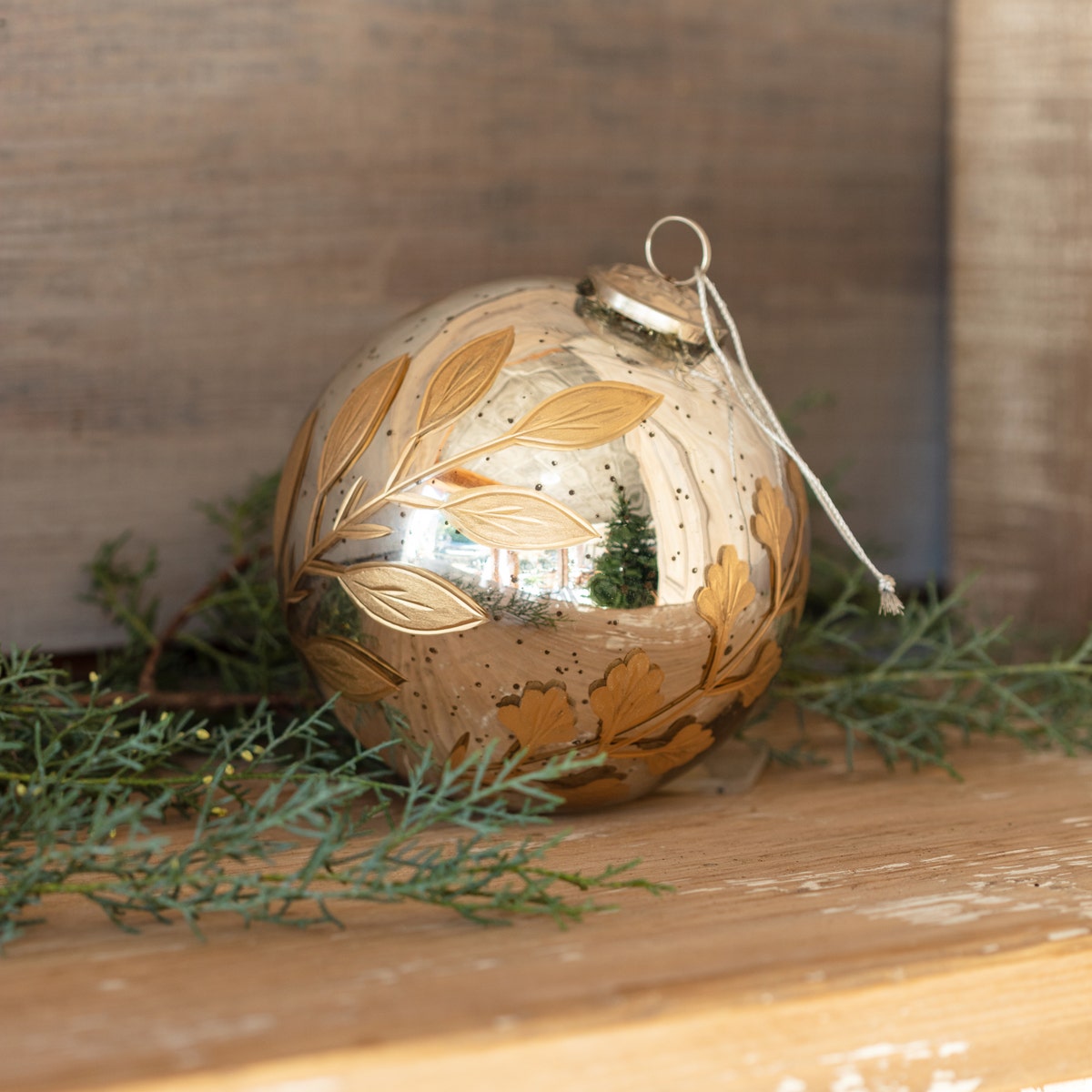 Round 6-inch antique mercury glass ornament with gold etched botanical leaf designs on a wood shelf.