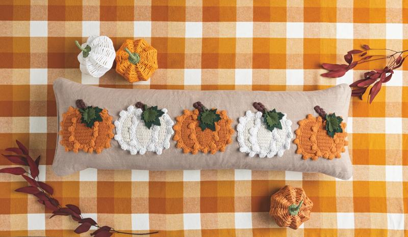 Long beige knit pillow featuring five orange and white textured pumpkin appliqués on a plaid cloth.