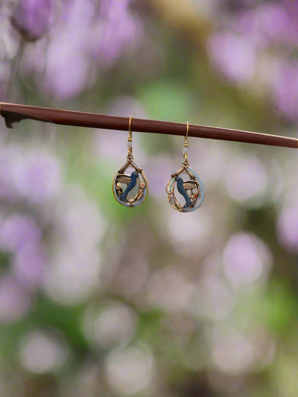 Hummingbird in Tear earrings with blue bird charms and brass frames hanging from a wooden branch.