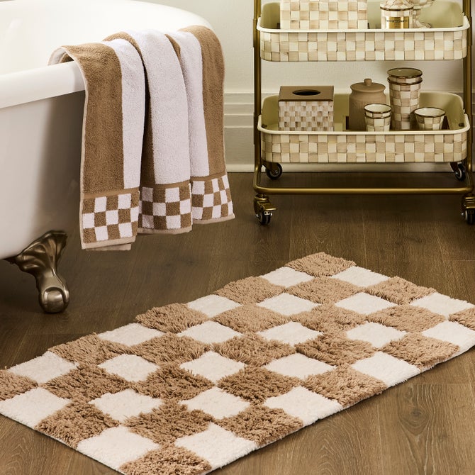 A rectangular checkerboard taupe and white bath rug on a wooden floor next to a clawfoot tub.