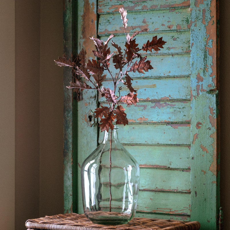 A metallic copper-colored oak leaf stem displayed in a clear glass bottle against a rustic shutter.
