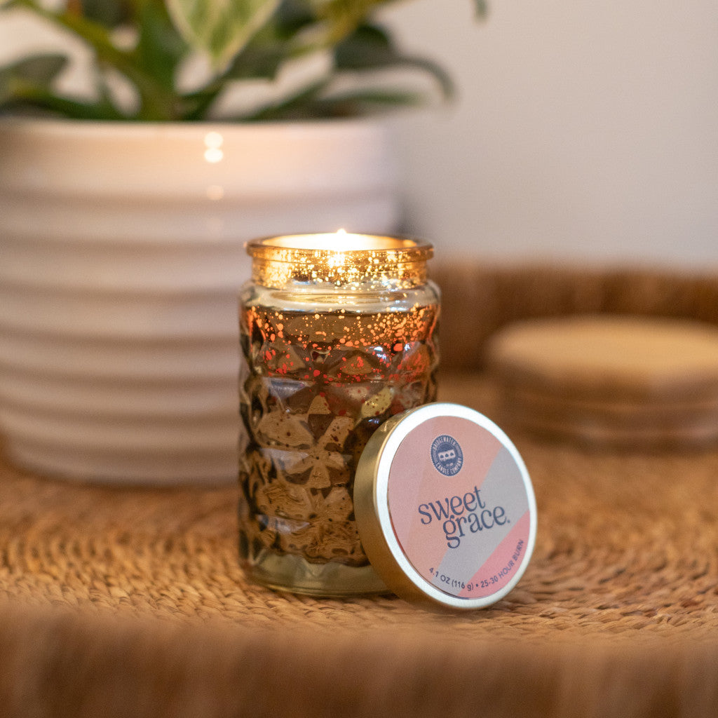 Sweet Grace scented candle in a textured glass jar with a lit wick and its lid resting beside it.