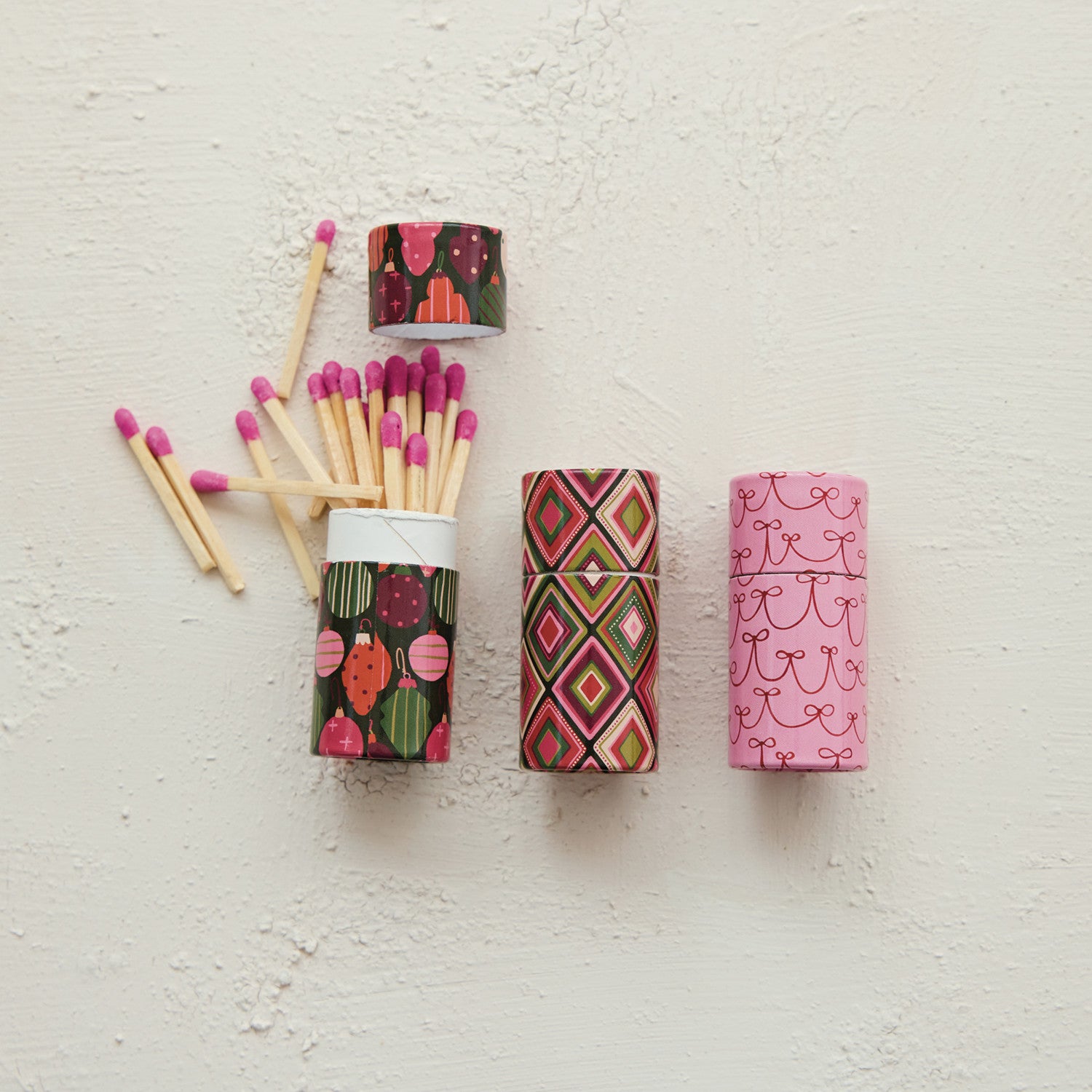 Three small tubes of safety matches with holiday patterns: ornaments, diamonds, and pink bows.