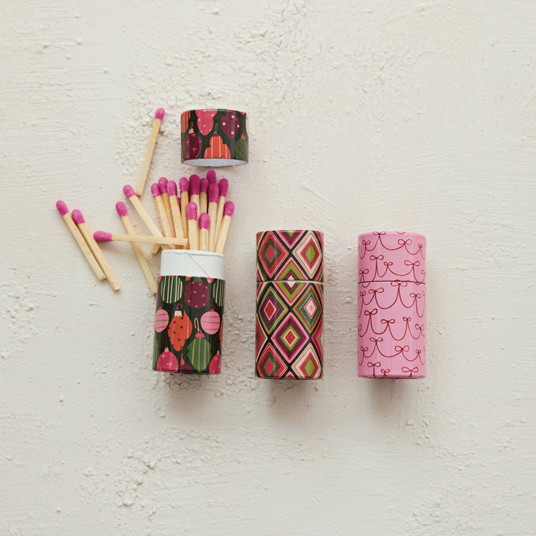 Three small tubes of safety matches with holiday patterns: ornaments, diamonds, and pink bows.