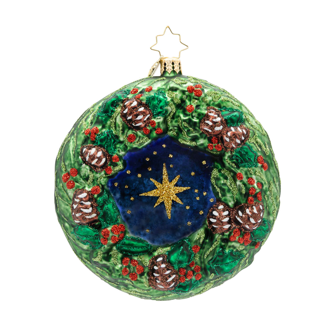 Round glass Christmas ornament featuring a green holly wreath with pinecones and a blue star center.