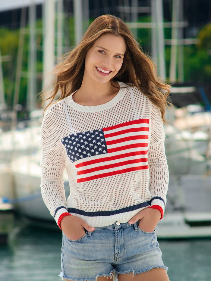 Woman wearing a white Cassie American Flag Mesh Sweater with denim shorts in a marina.