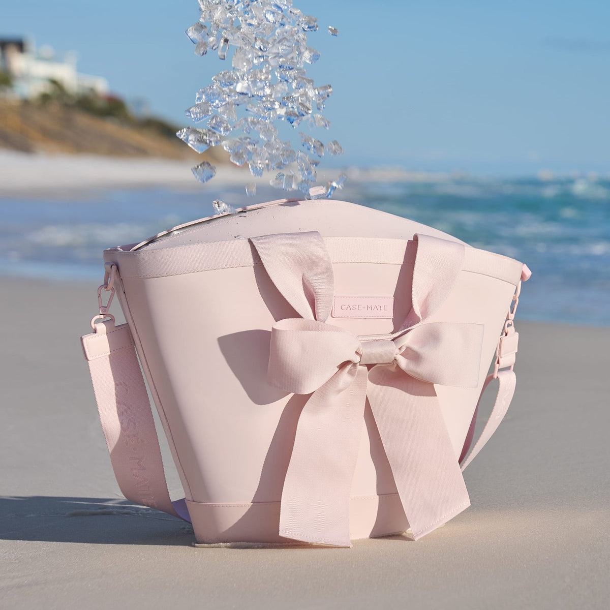 Ballet pink jelly cooler tote with a large bow on the front, sitting on a beach with ice falling in.
