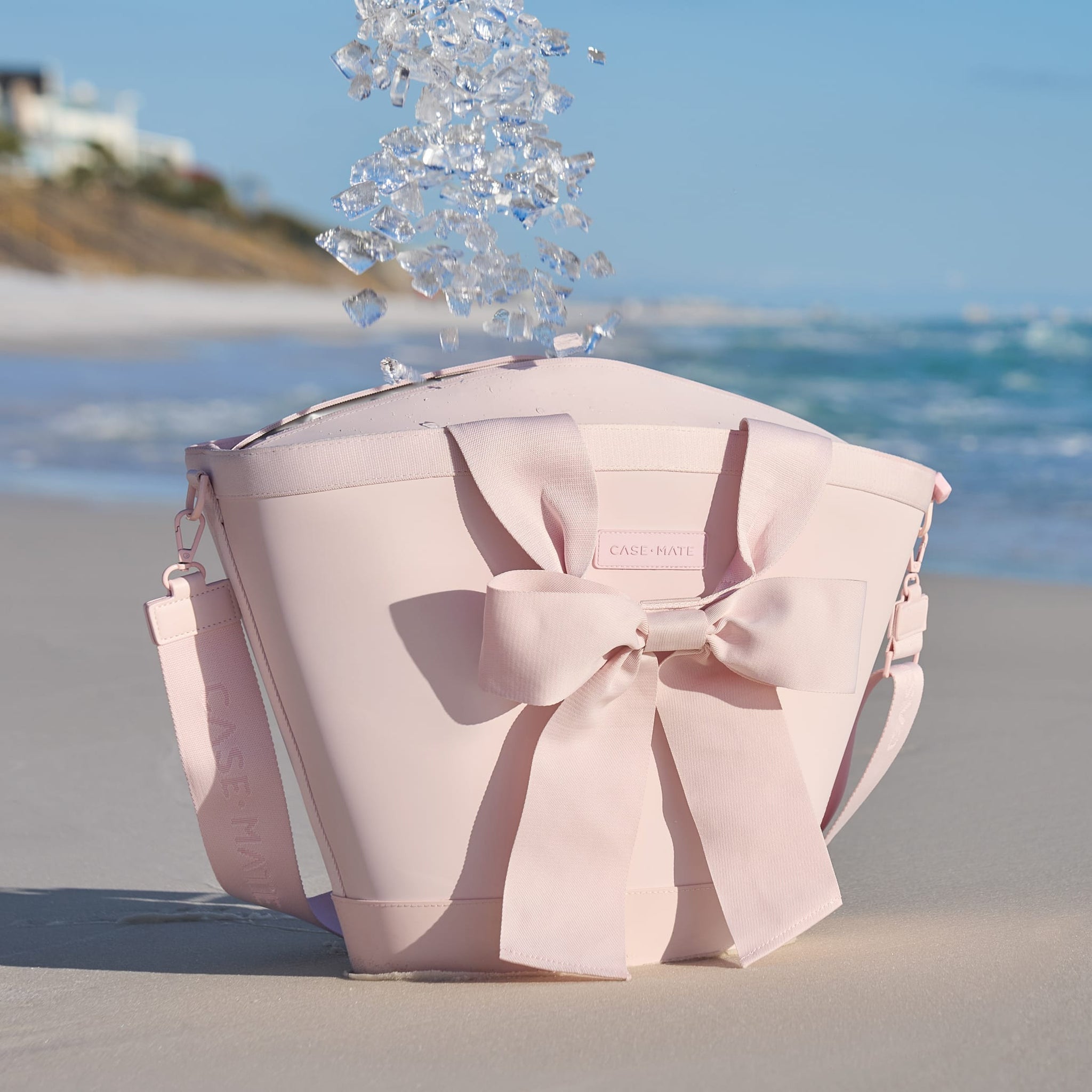 Ballet pink jelly cooler tote with a large bow on the front, sitting on a beach with ice falling in.
