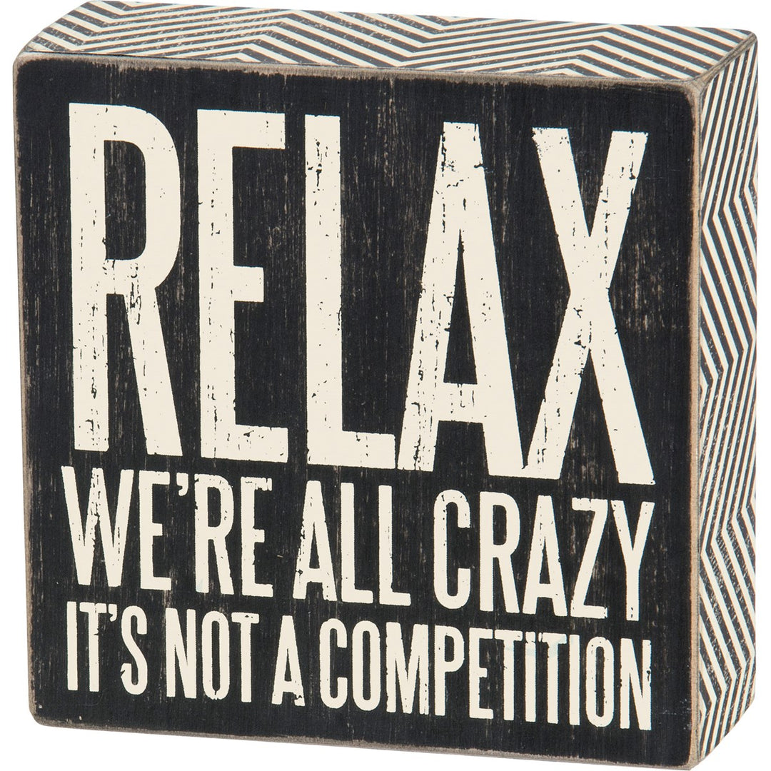 Black wooden box sign with white distressed text reading "RELAX WE'RE ALL CRAZY IT'S NOT A COMPETITION".