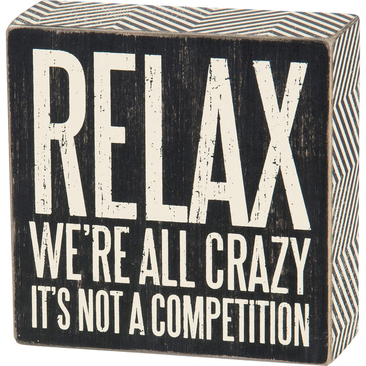 Black wooden box sign with white distressed text reading "RELAX WE'RE ALL CRAZY IT'S NOT A COMPETITION".