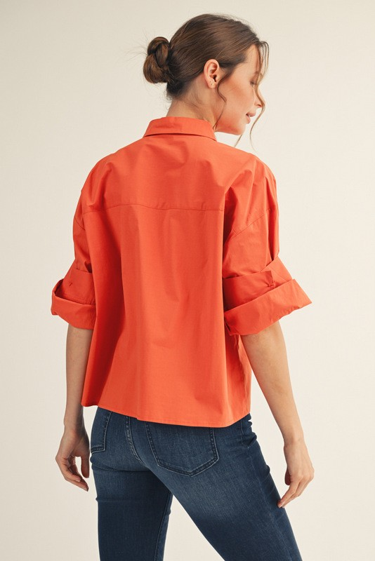 Back view of a woman wearing an orange Clarisse button-down shirt with multi-fold sleeves and jeans.