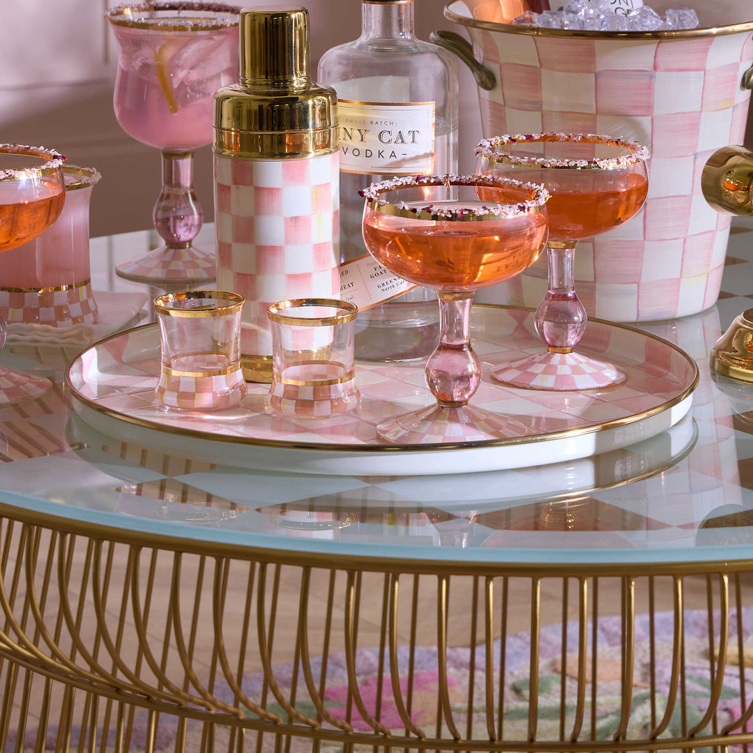A Rosy Check Round Tray on a glass table holding pink cocktails, shot glasses, and a shaker.