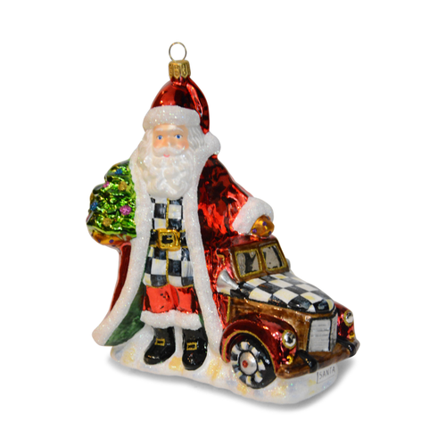 Glass Santa ornament in a red coat standing next to a vintage red truck with a checkered hood.