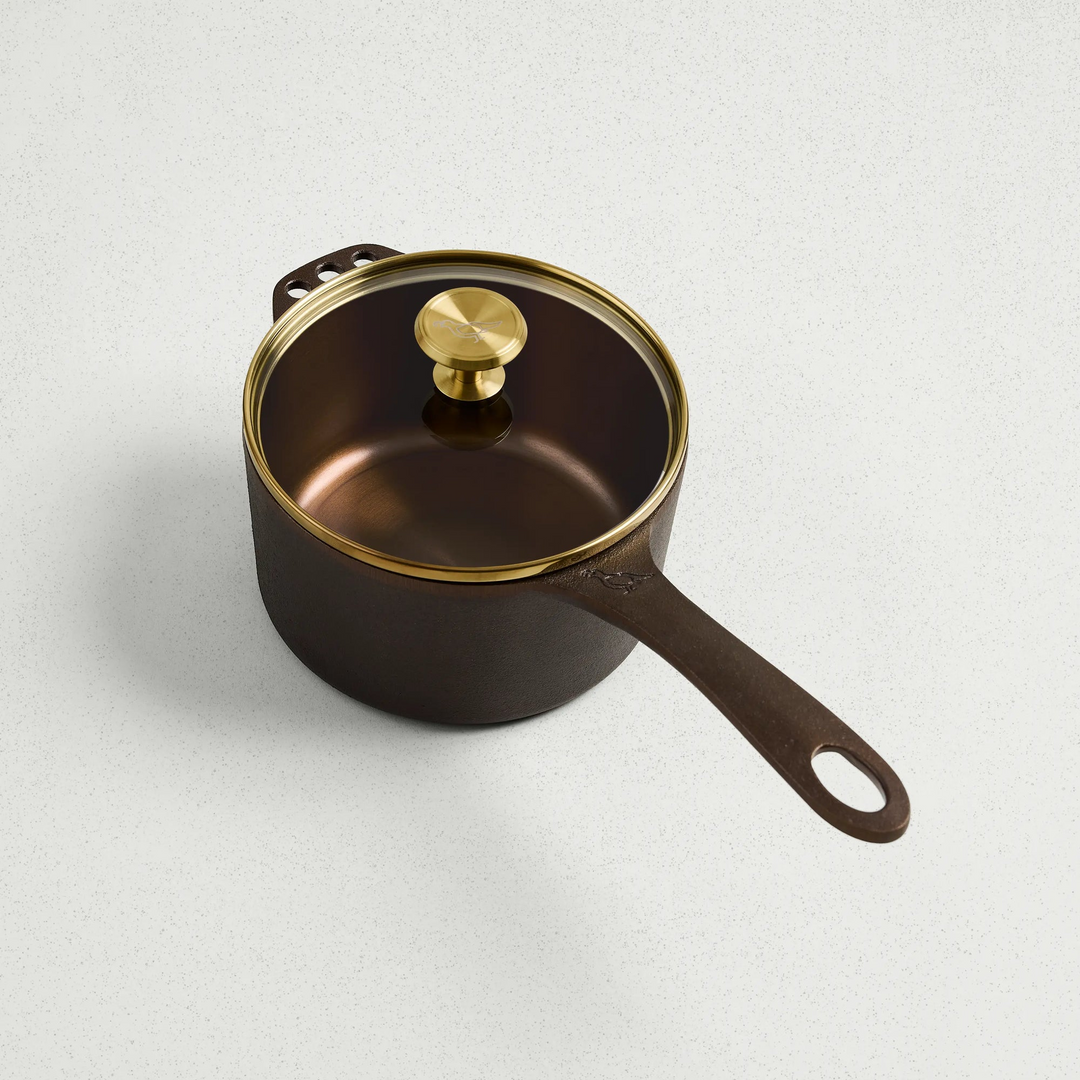 1 Qt brown cast iron saucepan with a glass lid and gold-toned knob on a white surface.