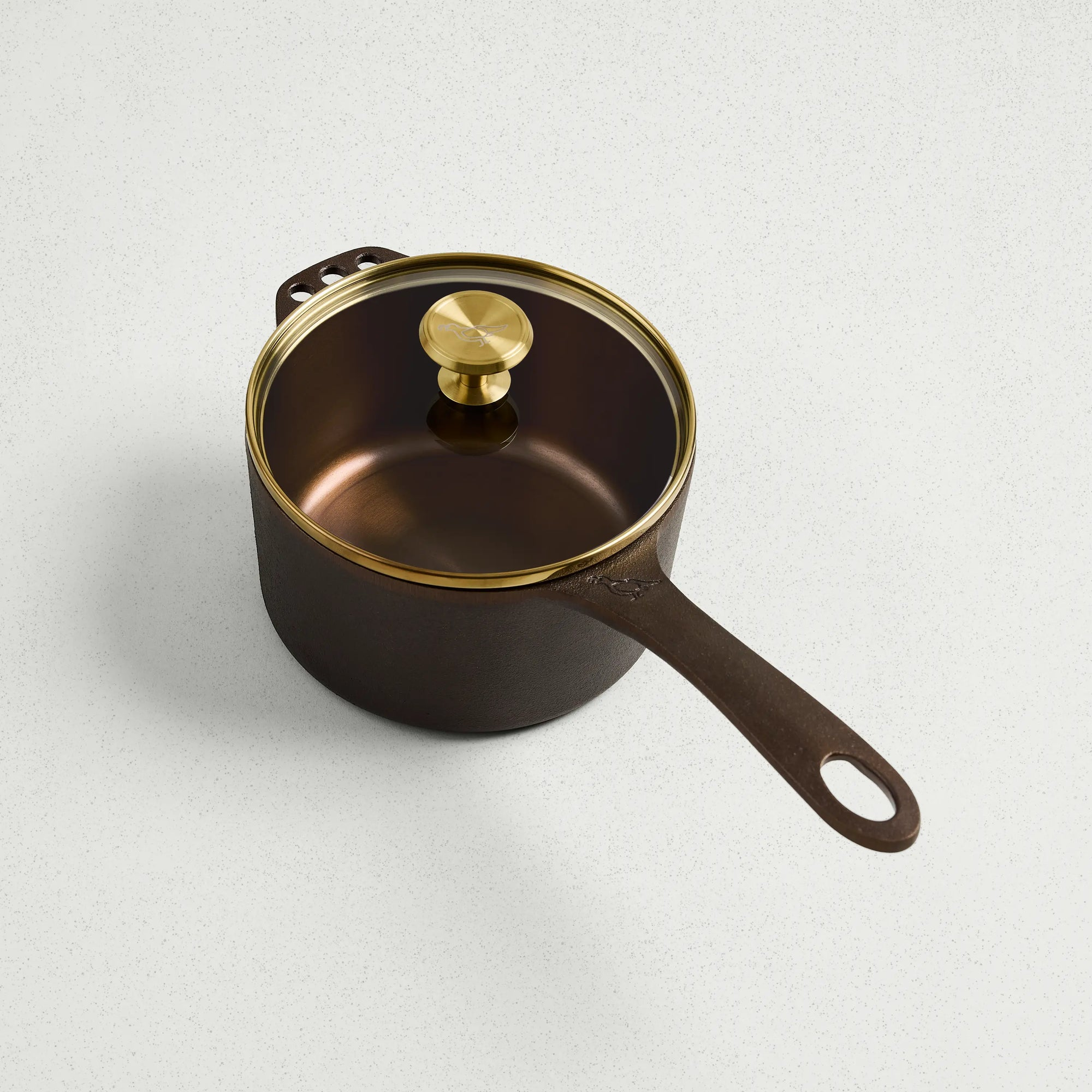 1 Qt brown cast iron saucepan with a glass lid and gold-toned knob on a white surface.
