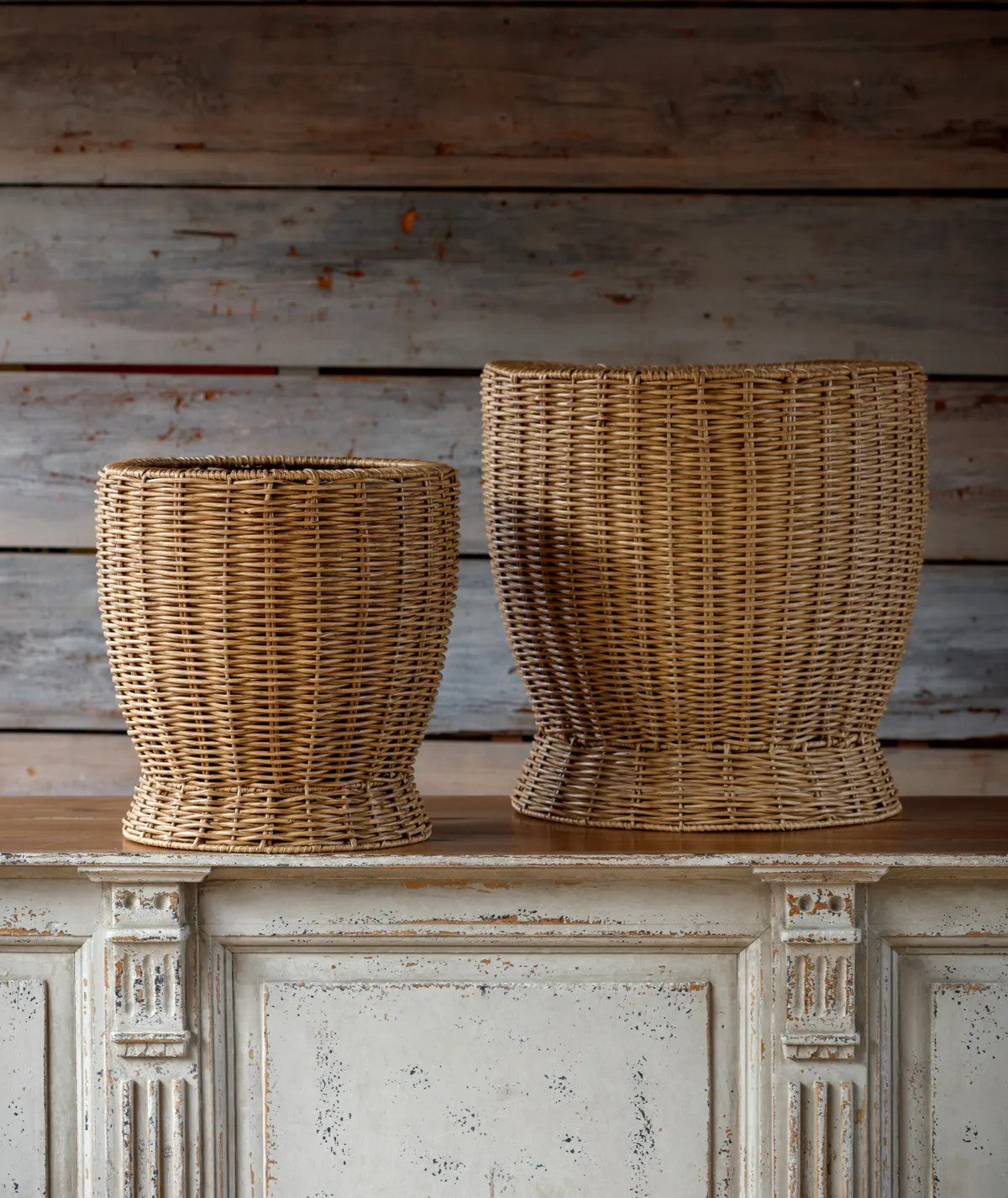 Two woven baskets, one smaller and one larger, made of natural-colored poly weave material, sit on a distressed white cabinet.