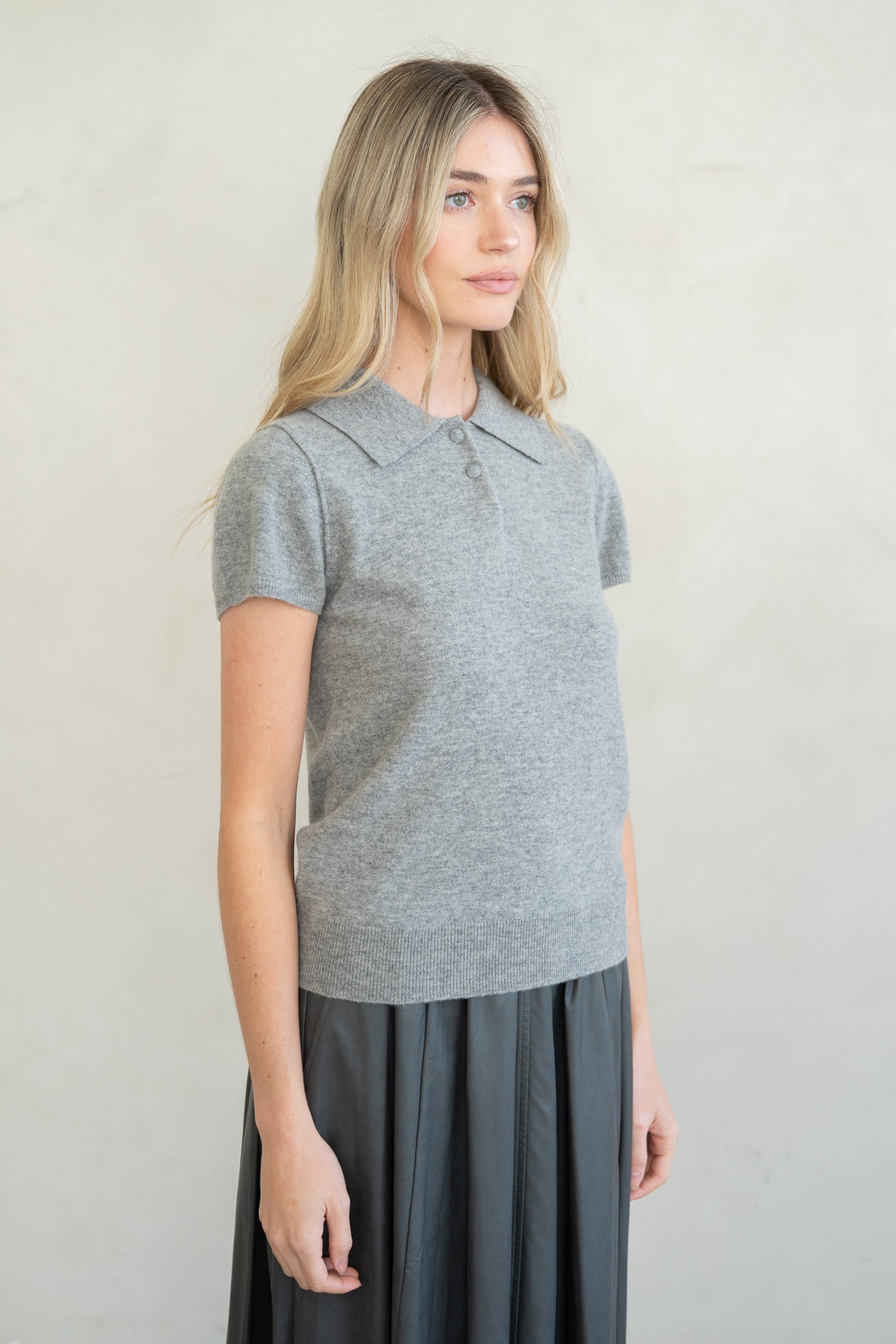 Woman wearing a grey Elise Refined Cap Sleeve Polo Sweater Top with a dark pleated skirt.