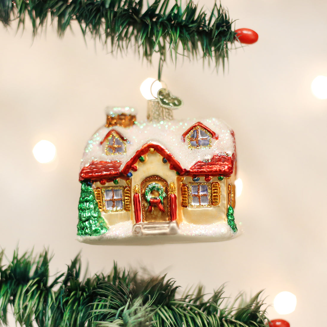 Glittery glass Holiday Home Ornament shaped like a snow-covered cottage hanging on a Christmas tree.