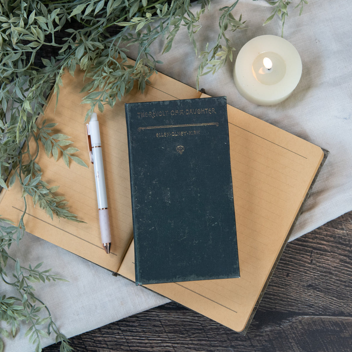 The Revolt of a Daughter journal with a dark green cover on an open notebook with a pen and candle.