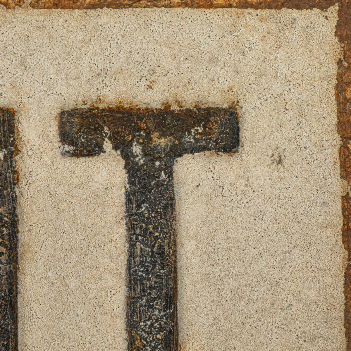 Close-up of the embossed letter T on a distressed metal wall sign with a textured beige background.