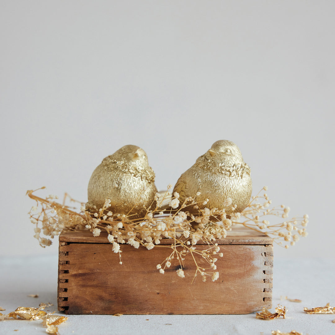 Two gold resin bird figurines with an artisan finish on a wooden box with dried flowers.