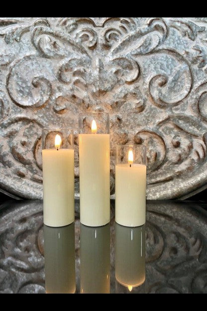 Three ivory pillar candles of varying heights in glass cylinders, set against a carved background.