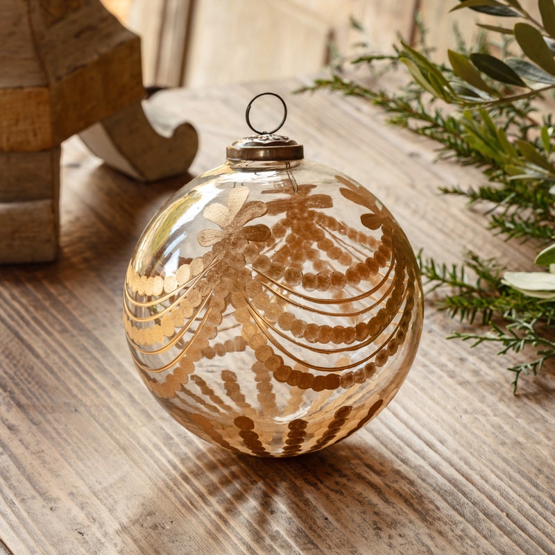 Clear glass ball ornament with etched and gilded golden garland design on a wooden surface.